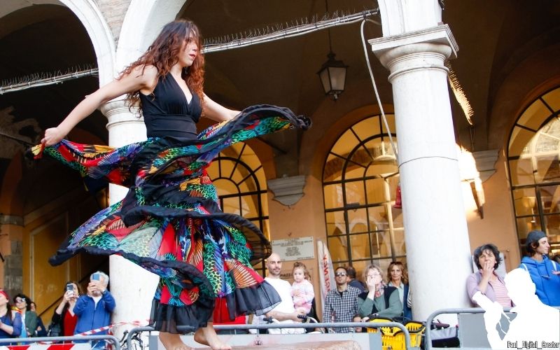 una ballerina con un vestito nero e con parti molto colorate e sgargianti che ondeggia sulla pietra Ringadora in Piazza Grande a Modena