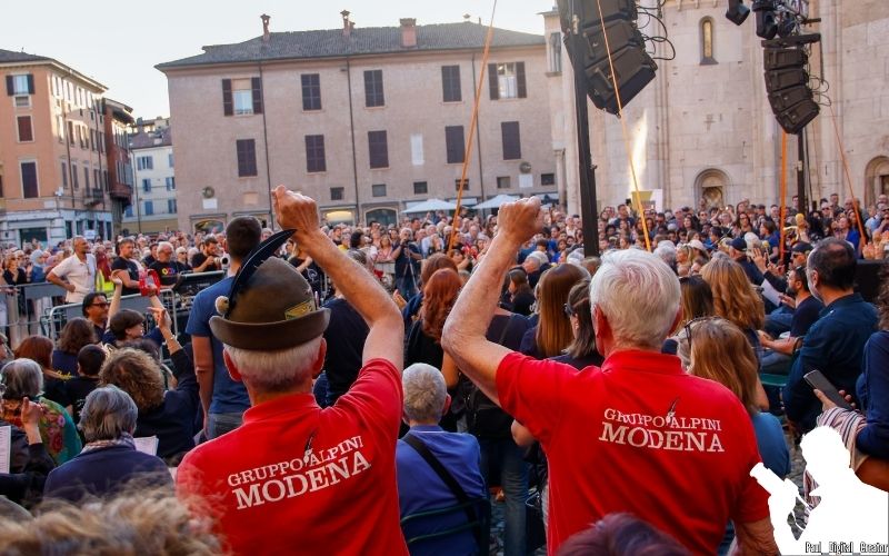 Due alpini che fanno il gesto della resistenza, uno dei due indossa il tipico cappello da Alpino, entrambi indossano maglie rosse e sono voltati verso il pubblico, visti di schiena nella foto
