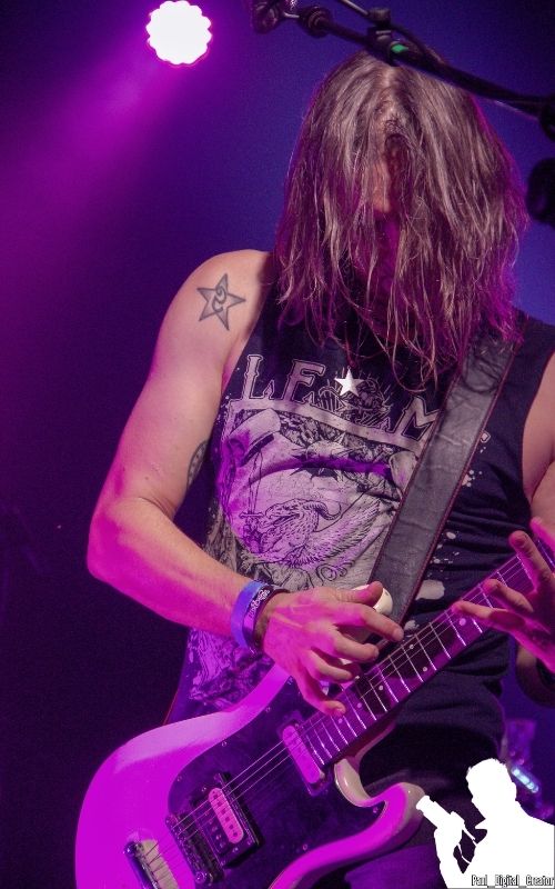 il chitarrista di Speedstroke eseguendo un assolo, ha la faccia quasi interamente ricoperta dai capelli, una maglia nera senza maniche ed è circondato da luci blu e viola e la chitarra bianca