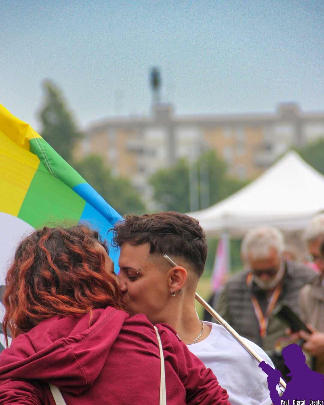 Un bacio tra due ragazze, una con capelli lunghi ed una con capelli corti la quale tiene in mano una bandiera di Arcigay.